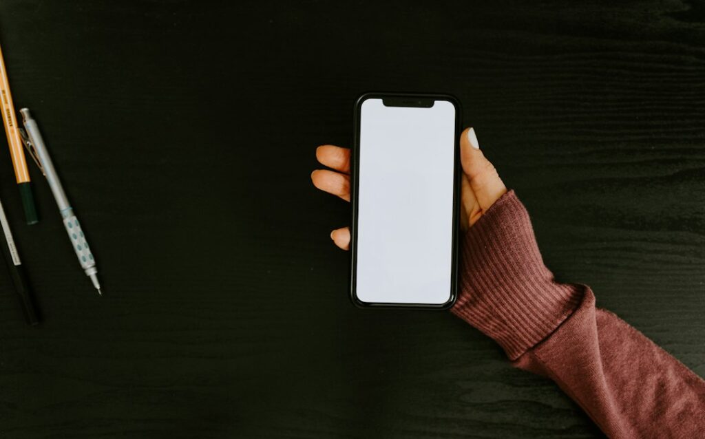 筆記具を置いた暗い木目の机で、白い画面のスマートフォンを持つ手元